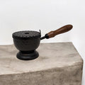 Crystal Fairy Patch 9-inch matte black hammered iron cauldron incense burner with a perforated lid and wooden handle on stone.