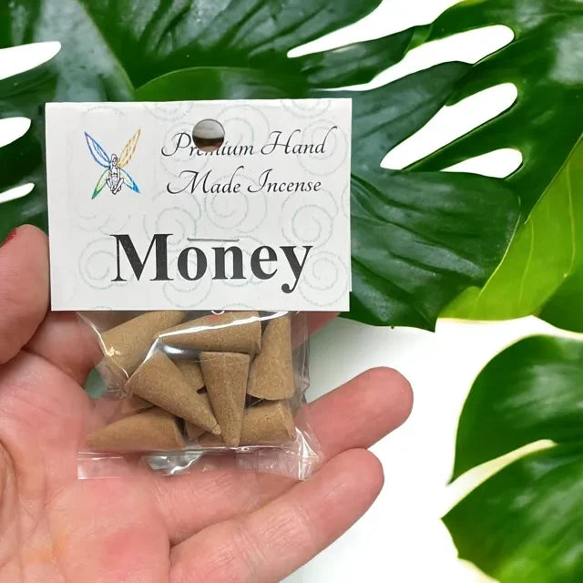 Crystal Fairy Patch Money premium hand-made incense cones in a clear plastic bag held against a green monstera leaf background.